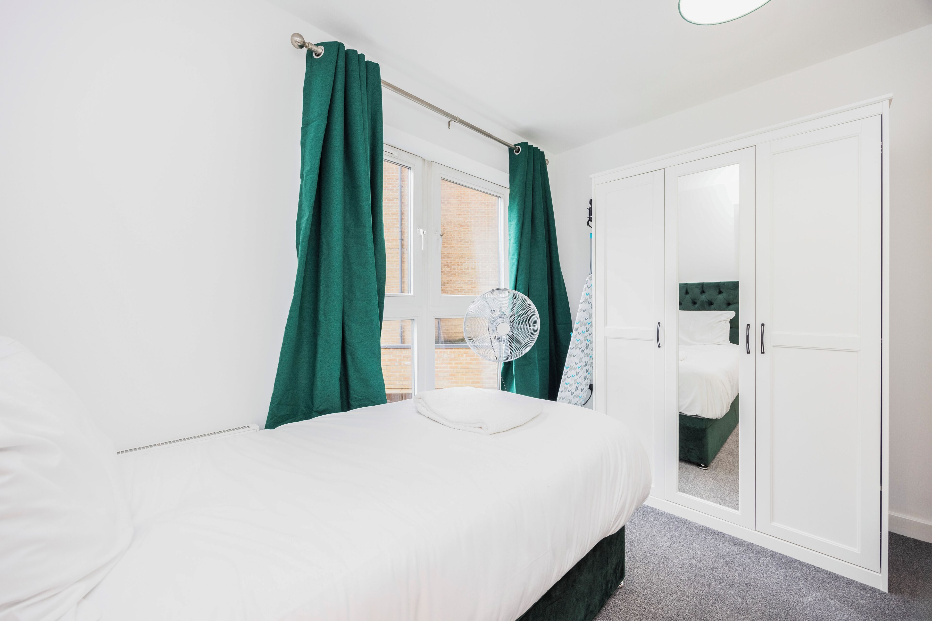A bedroom with a bed, a fan, a wardrobe, and a window with curtains.