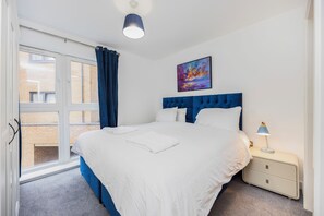 A bedroom with a large bed, a nightstand with a lamp, a window with curtains, and a painting on the wall. - Charming Apartment for Friends, Couples and Families in London - Hackney 3 (London)