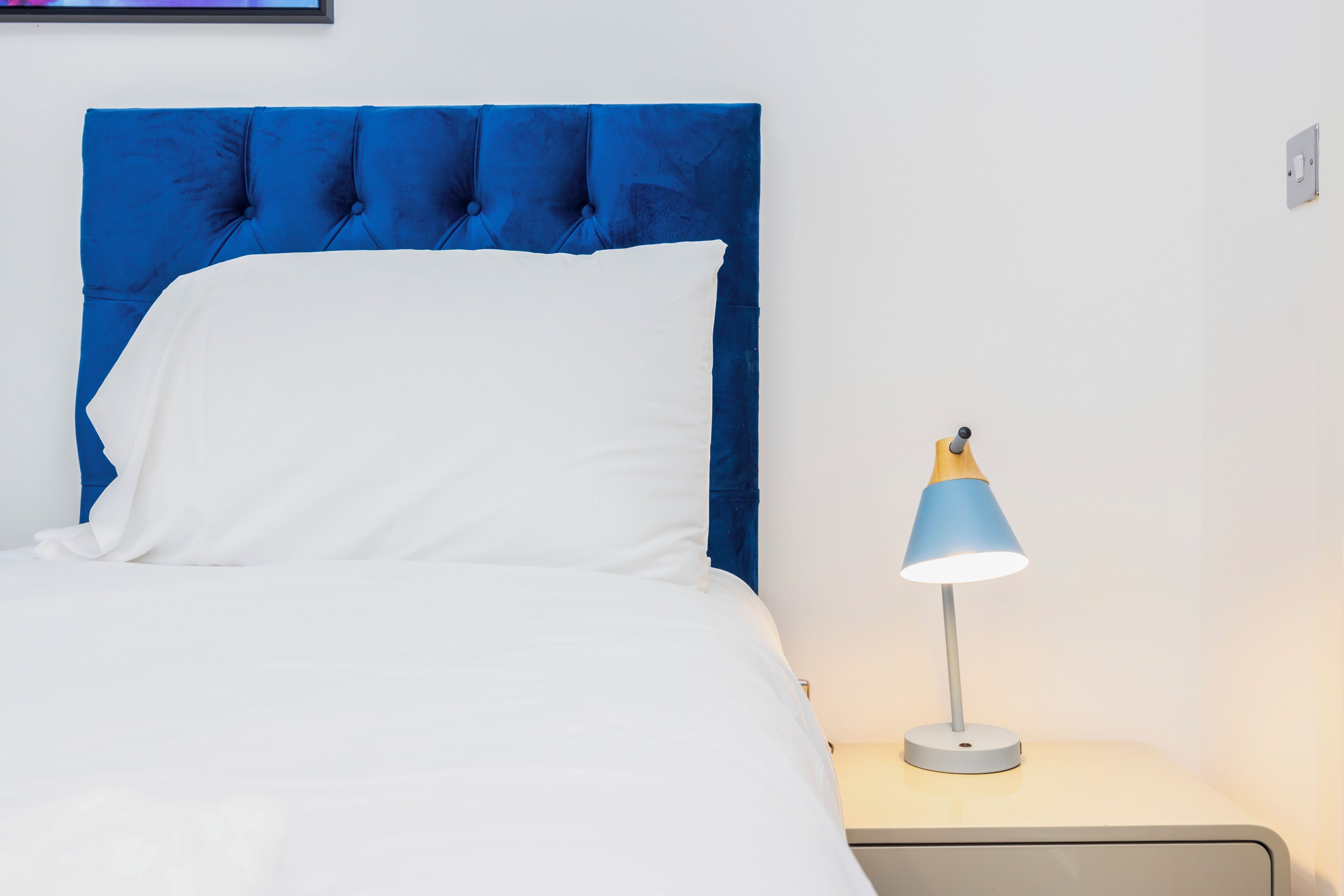 A bed with a blue headboard, a white pillow, and a bedside table with a lamp.