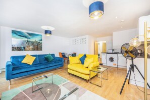 A modern living room with a blue sofa, a yellow sofa, a glass coffee table, and a kitchen area in the background. - Charming Apartment for Friends, Couples and Families in London - Hackney 3 (London)
