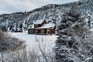 A wooden cabin with a metal roof, surrounded by snow-covered trees and a hill in the background. - Mountain view modern home with open layout, gas fireplace, smart TVs, & W/D (South Fork)