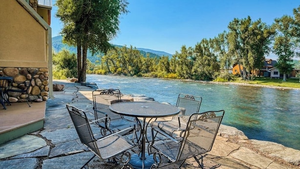 Une terrasse avec une table ronde et des chaises donnant sur une rivière, entourée d’arbres et d’un mur de pierre.