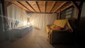 Een kamer met een houten plafond, een bank met kussens, een staande ventilator en een tafel met een lamp.