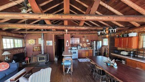 Ein rustikales Ferienhäuschen im Inneren mit Holzbalkendecke, Küchenbereich, Esstisch und Wohnzimmer mit Sofa und Fernseher.