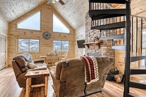 Ein gemütliches Blockhaus-Interieur mit Holzausstattung, einem Kamin aus Stein, einer Wendeltreppe und großen Fenstern mit Blick auf die verschneite Landschaft.