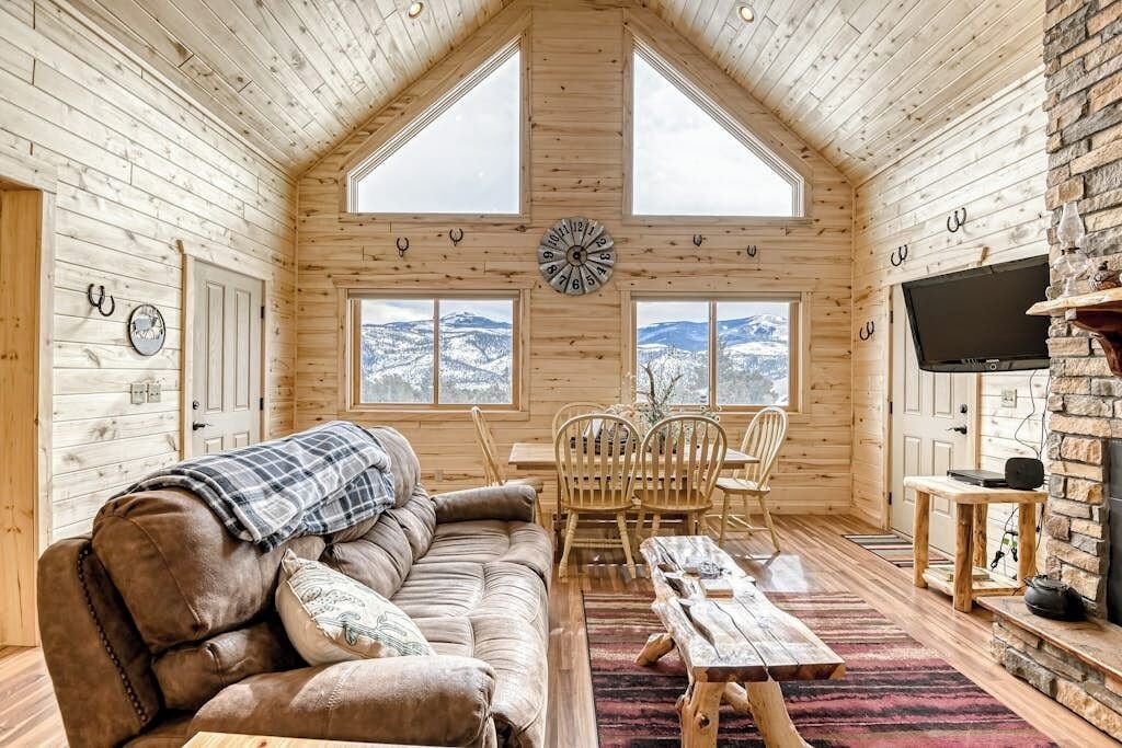 A cozy cabin interior with a wooden ceiling, a large window, a fireplace, and a television.