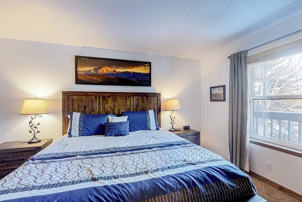 Un dormitorio con cabecera de madera, cama con ropa de cama azul, dos lámparas de noche, un cuadro sobre la cama y una ventana con cortinas.