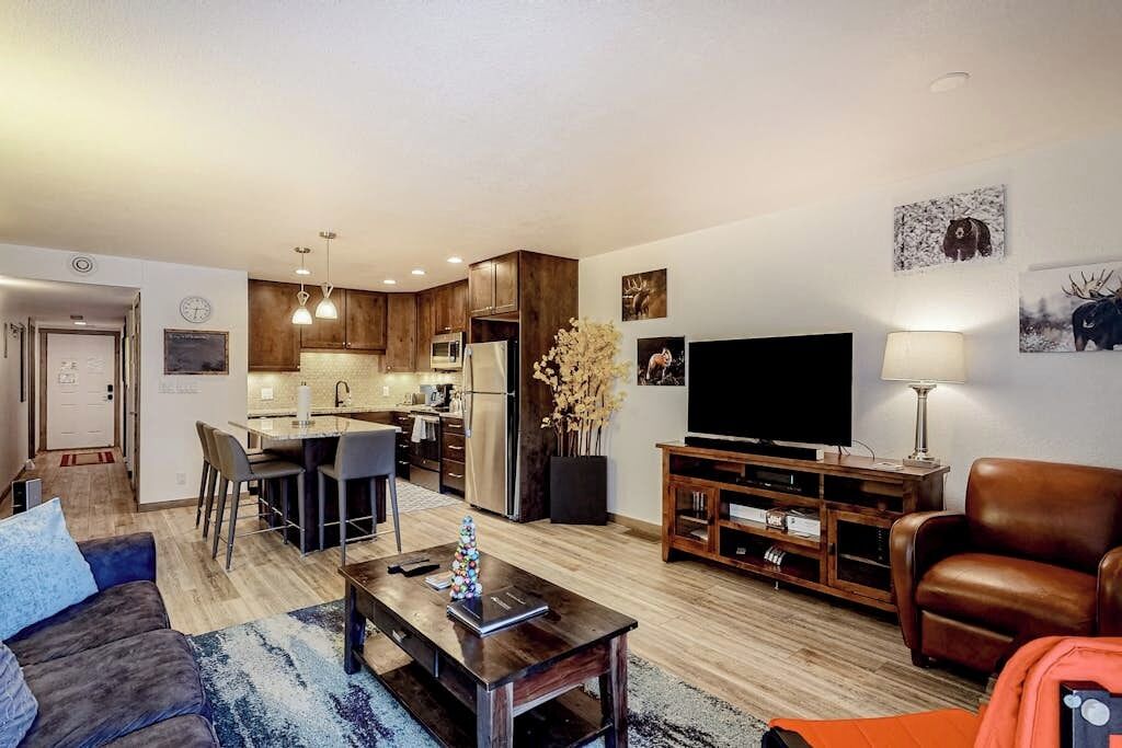 Una sala de estar moderna con un sofá, una mesa de centro, un televisor y un comedor con sillas, además de una cocina al fondo.