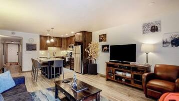 Una sala de estar moderna con un sofá, una mesa de centro, un televisor y un comedor con sillas, además de una cocina al fondo.