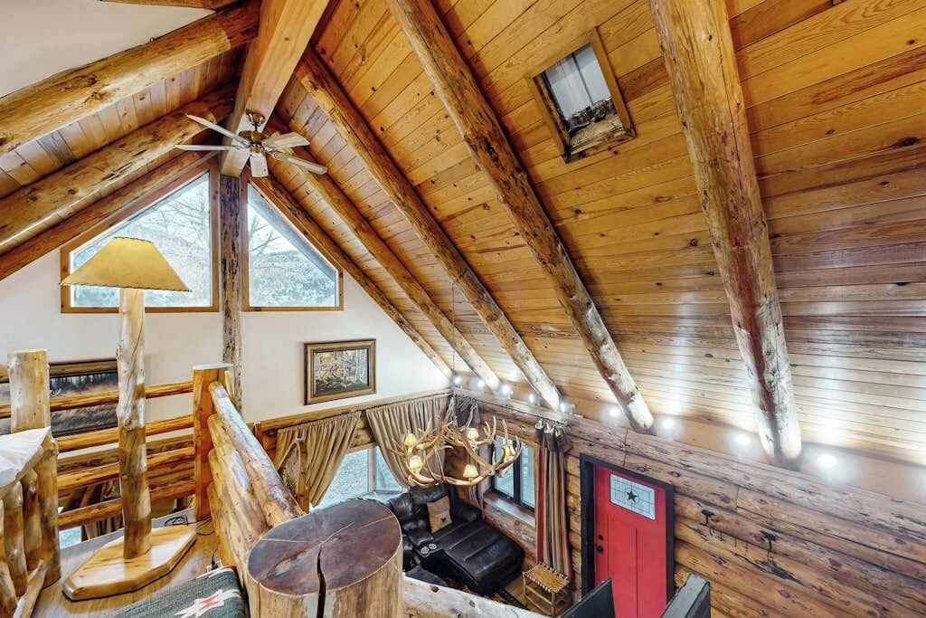 L’intérieur d’une cabane en rondins, avec un plafond en pente, un ventilateur de plafond, une cheminée, un canapé et une table à manger.