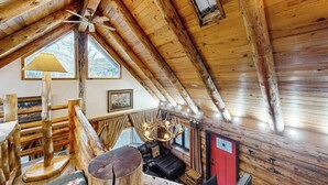 A log cabin interior with a sloped ceiling, a ceiling fan, a fireplace, a sofa, and a dining table.
