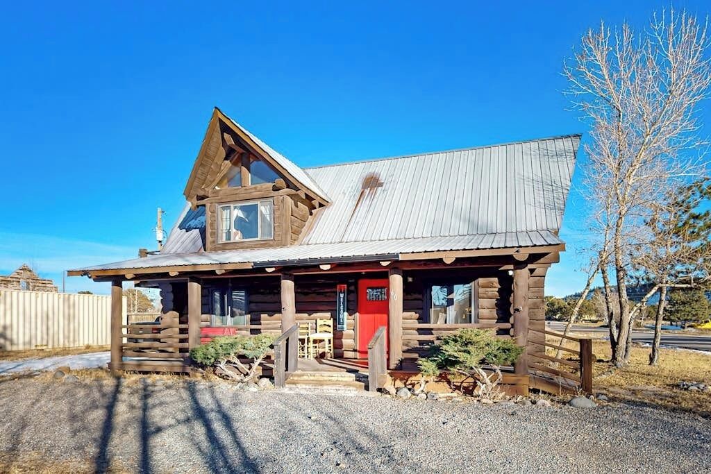 Une cabane rustique en rondins de bois, avec un toit en métal, une véranda et une porte rouge.