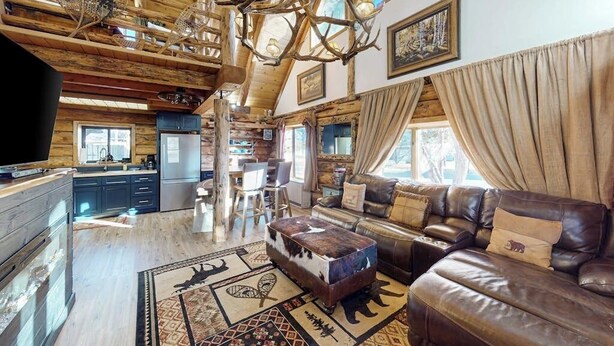 A rustic cabin interior with a wooden ceiling, a leather sofa, a dining area, and a kitchen.
