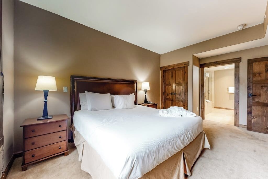 Une chambre à coucher avec un grand lit, deux tables de chevet avec des lampes, une armoire en bois et une porte donnant sur une autre pièce.