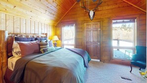 A wooden cabin bedroom with a bed, a ceiling fan, a mounted deer skull, and a view of the outdoors. - Riverfront home with river & valley views, wood stove, deck, & W/D (South Fork)