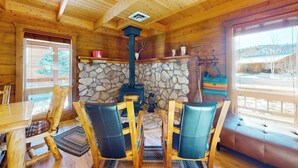 A wooden cabin interior with a stone fireplace, wooden chairs, a bench, and a window with a view. - Riverfront home with river & valley views, wood stove, deck, & W/D (South Fork)