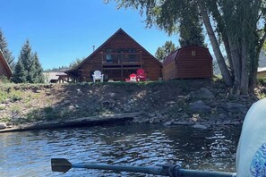 Eine Hütte mit Terrasse, zwei Adirondack-Stühlen und einem Schuppen an einem Fluss.