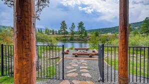 Une porte en bois rustique qui mène à un sentier pavé de pierres, bordée d’une clôture en métal, avec une rivière et des arbres en arrière-plan.
