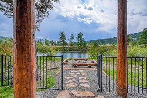 Une porte en bois rustique qui mène à un sentier pavé de pierres, bordée d’une clôture en métal, avec une rivière et des arbres en arrière-plan.