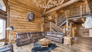 L’intérieur d’une cabane en rondins, avec un canapé d’angle en cuir, une table basse en bois, un tapis en cuir noir, un lustre et un escalier.