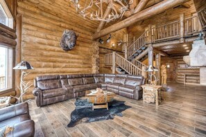 L’intérieur d’une cabane en rondins, avec un canapé d’angle en cuir, une table basse en bois, un tapis en cuir noir, un lustre et un escalier.