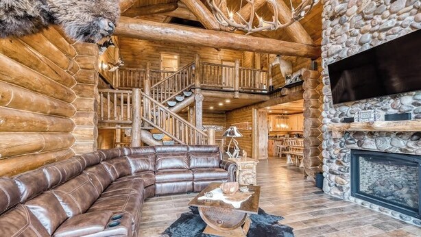 L’intérieur d’une cabane en rondins, avec un canapé en cuir, une cheminée en pierre et un escalier.