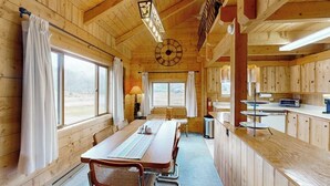Interior de uma cabana de madeira com mesa de jantar, cadeiras, cozinha e janelas amplas.