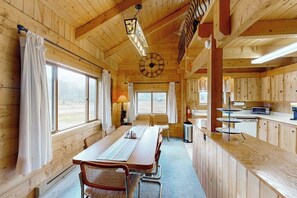 Ein Interieur einer Holzhütte mit Esstisch, Stühlen, einer Küchenzeile und großen Fenstern.