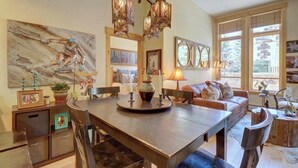 Un coin repas avec une table en bois, des chaises et un lustre. Les murs sont ornés de tableaux encadrés, et il y a un canapé avec des coussins.