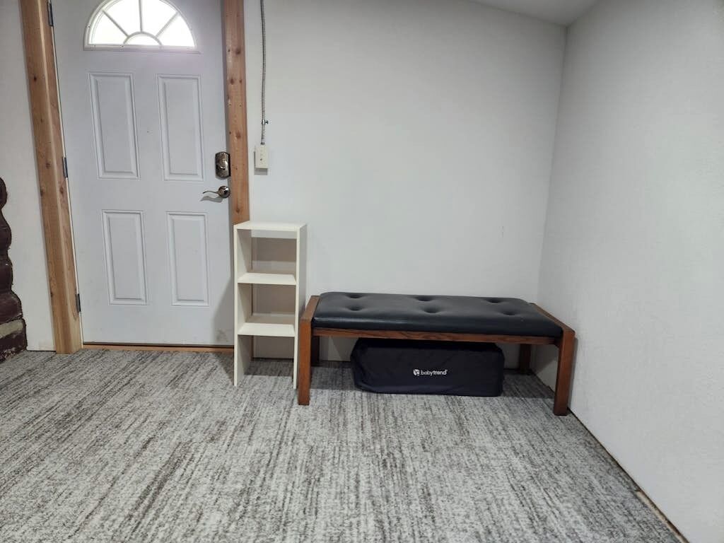 Ein Zimmer mit einer weißen Tür, einer Bank, einem weißen Regal und einer schwarzen Tasche mit dem Aufdruck 'Babyfriend' auf dem Boden.