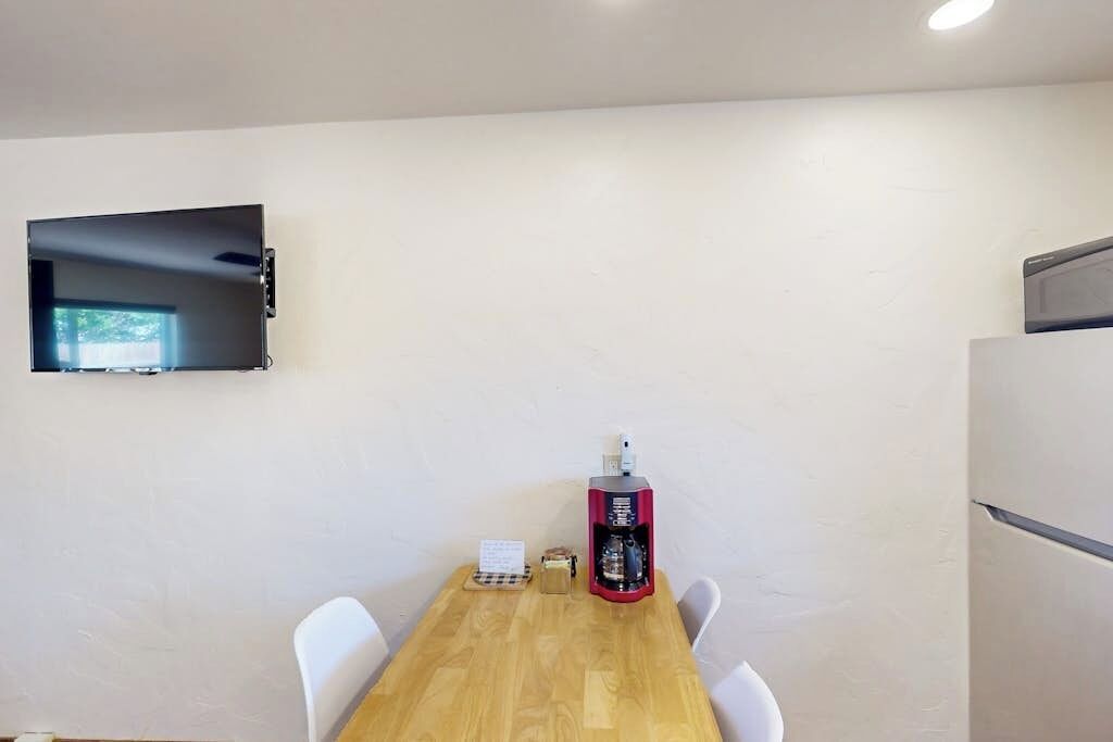 Une pièce avec une table en bois, des chaises blanches, un téléviseur à écran plat fixé au mur et une machine à café.