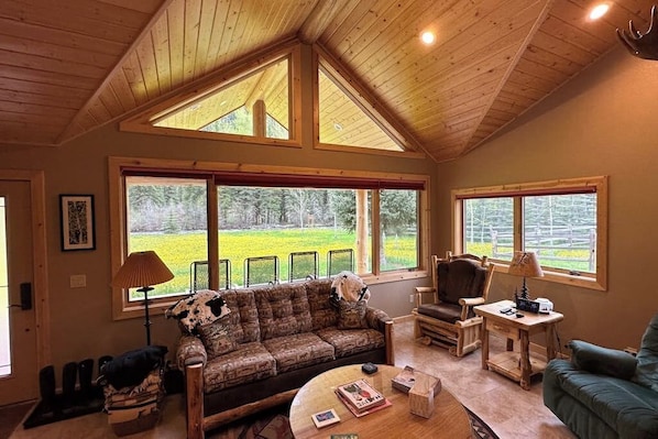 Ein gemütliches Wohnzimmer mit hoher Holzbalkendecke, großen Fenstern, einem gemusterten Sofa, einem Sessel, einem kleinen Tisch und einer Lampe.