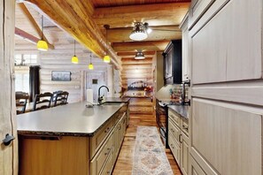 A kitchen with wooden beams, a central island, pendant lights, and a view into the dining area. - Charming cabin surrounded by trees with deck, grill, fireplace, games, & W/D (South Fork)