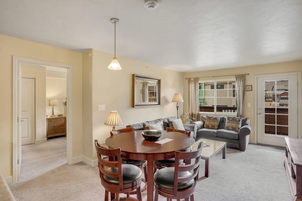 A living room with a sofa, a coffee table, and a dining table with chairs. There is a window with curtains, a door, and a mirror on the wall.