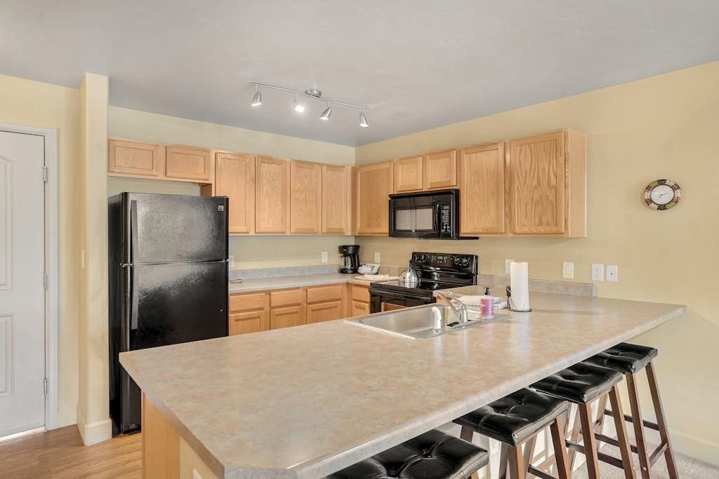 A kitchen with wooden cabinets, a black refrigerator, a microwave, a stove, a sink, and bar stools.