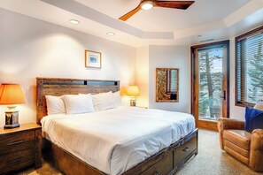 Ein Schlafzimmer mit einem großen Bett, einem hölzernen Kopfteil, zwei Nachttischlampen, einem Fenster mit Jalousien und Blick auf Bäume.