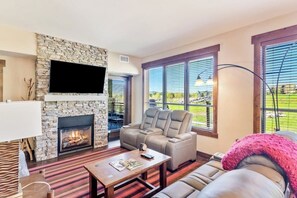 A living room with a stone fireplace, a flat-screen TV, a sectional sofa, and large windows with a view of a green landscape. - Condo with two balconies, fireplace, on-site gondola, pool & three hot tubs (Steamboat Springs)