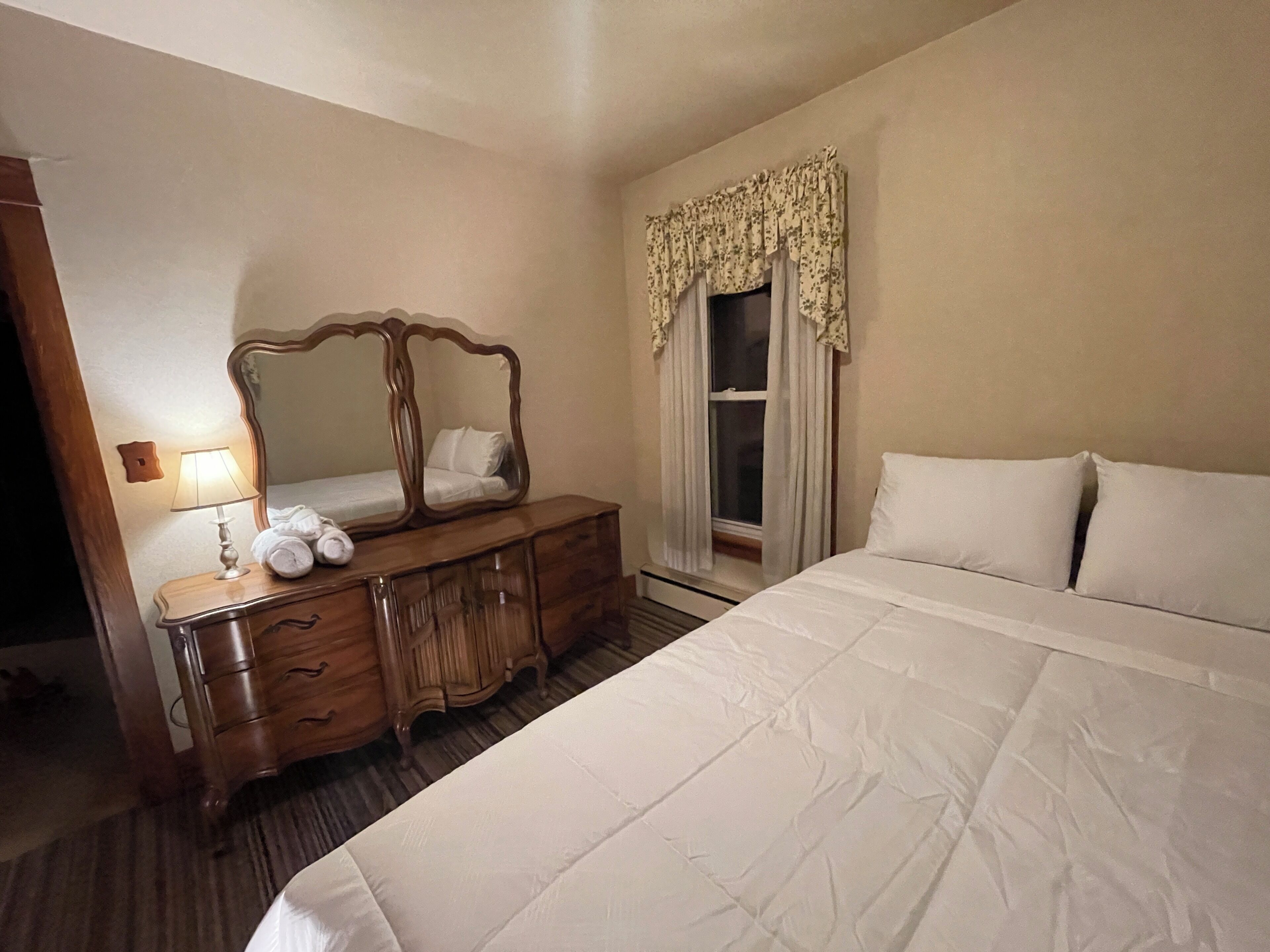 Une chambre d’hôtel avec un grand lit, une commode en bois avec un miroir, une fenêtre avec des rideaux et une lampe de chevet.