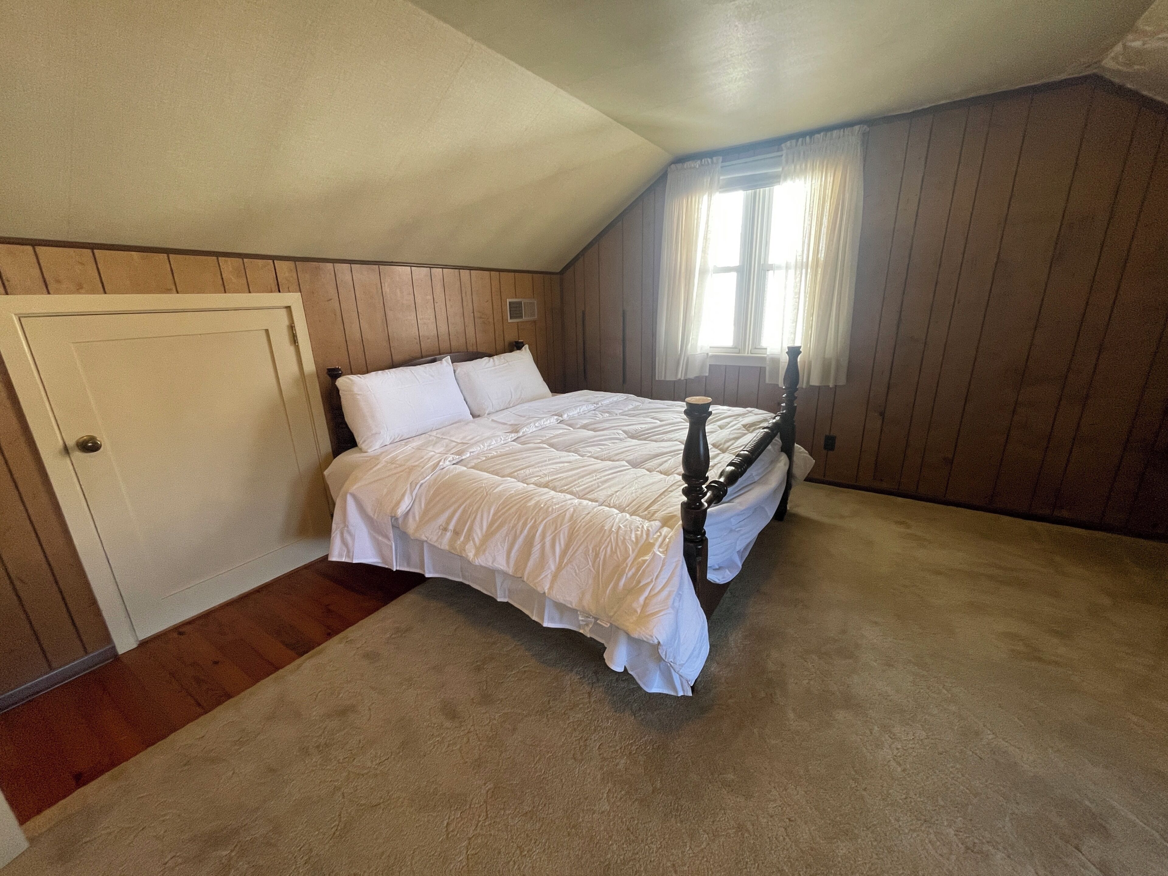 A bedroom with a single bed, wooden walls, a window with curtains, and a door.