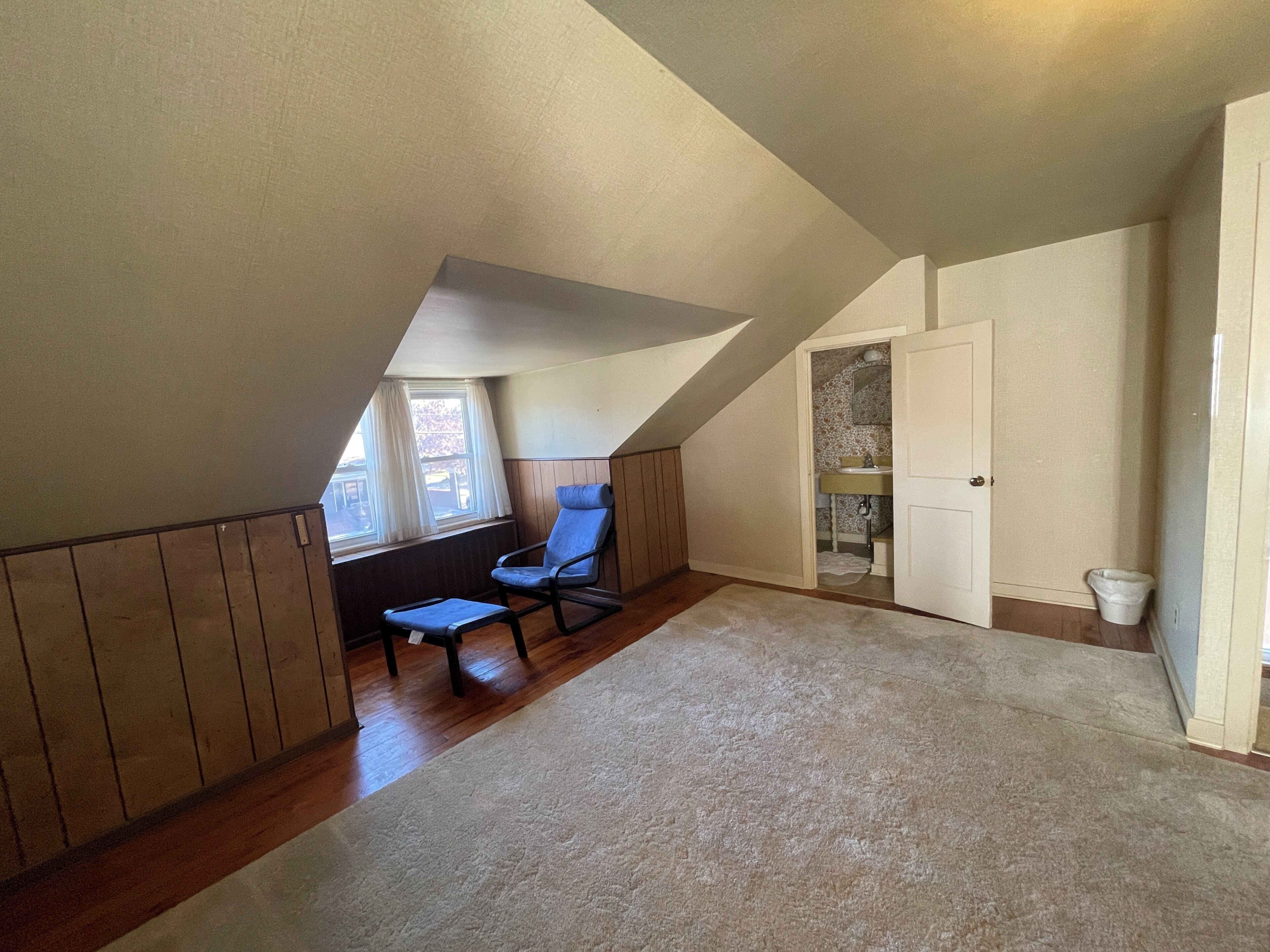 A room with a sloped ceiling, a rocking chair, a small window, and a door leading to a bathroom.