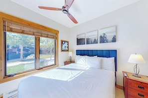 Ein Schlafzimmer mit Bett, Deckenventilator, Nachttisch, Lampe, Fenster mit Jalousien und Wandbildern.