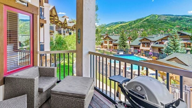 Un balcón con mobiliario de mimbre, una parrilla y vistas a un complejo turístico de montaña con casas y una piscina.