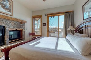 A hotel room with a stone fireplace, a bed, a TV, a balcony with a view, and a wooden headboard. - Top-floor, walk-to-lift, corner condo w/ fireplace, views & resort hot tubs/pool (Steamboat Springs)