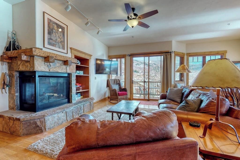 A living room with a fireplace, a ceiling fan, a sofa, a TV, and a large window.