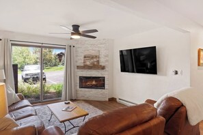 Ein Wohnzimmer mit Kamin, Deckenventilator, Flachbildfernseher und großem Fenster mit Blick auf ein Fahrzeug draußen.