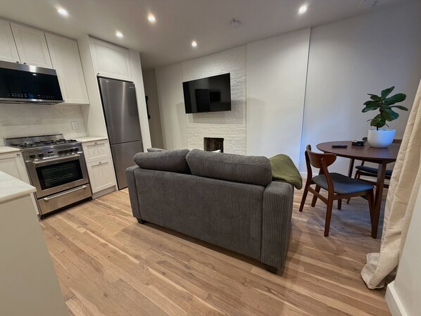 A modern kitchen with built-in appliances, a grey sofa, a dining table with chairs, and a flat-screen TV mounted on the wall.