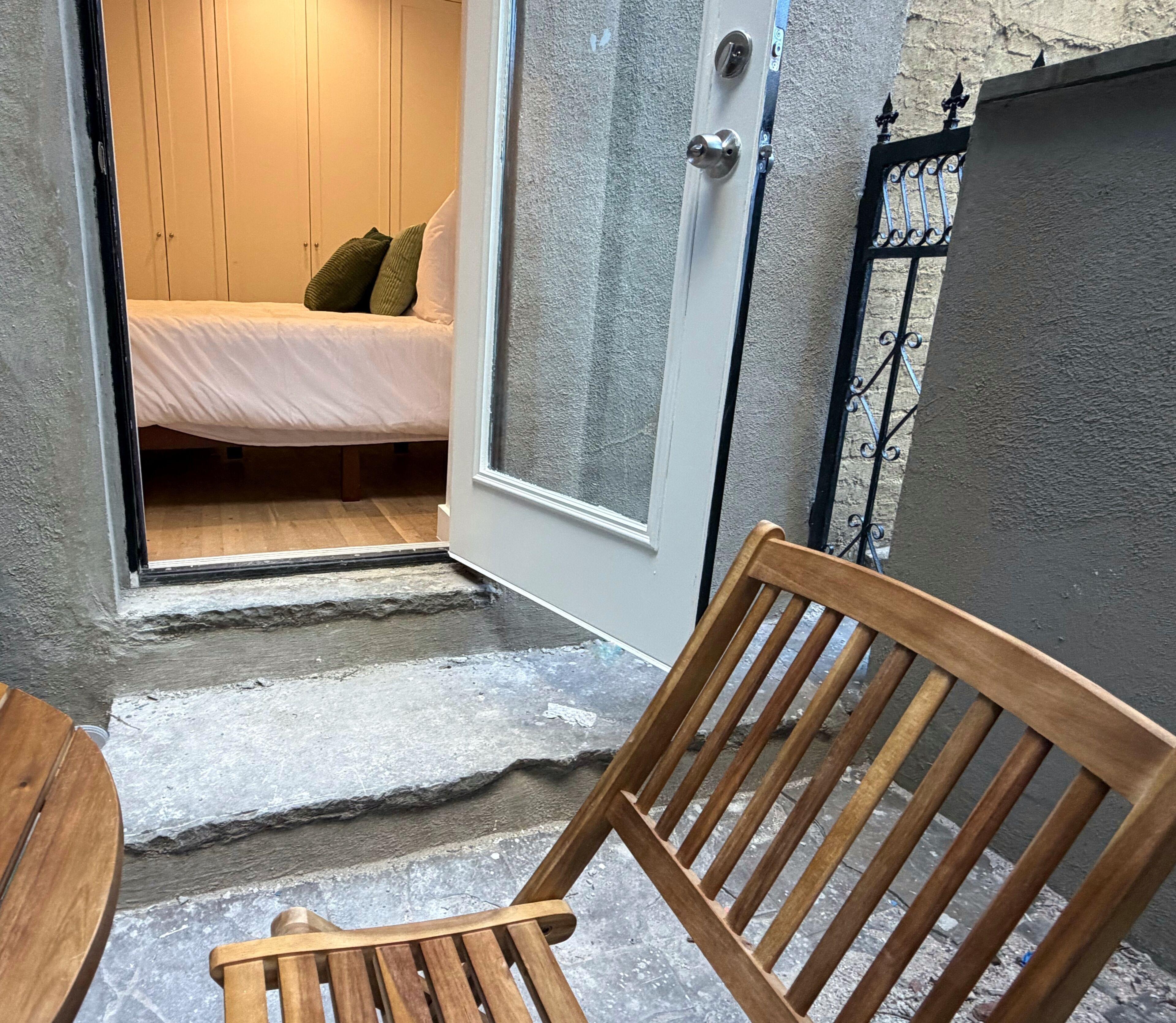 Une chaise en bois est placée dans l’embrasure d’une porte qui donne sur une pièce où se trouve un lit et une porte blanche.