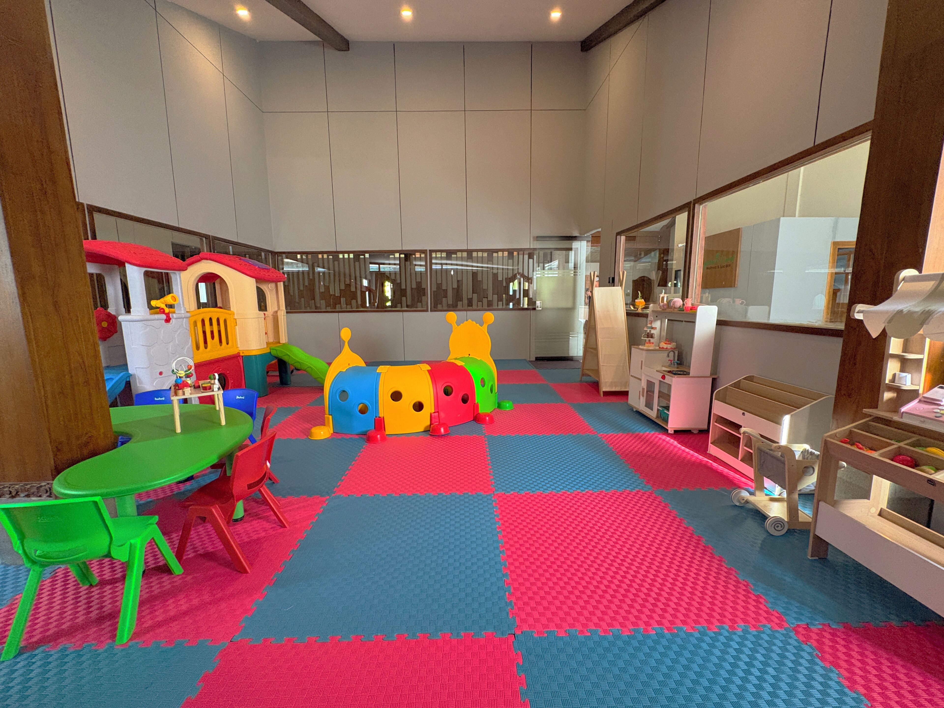 A children's play area with colorful toys and a playhouse, a table with chairs, and a mat-covered floor.