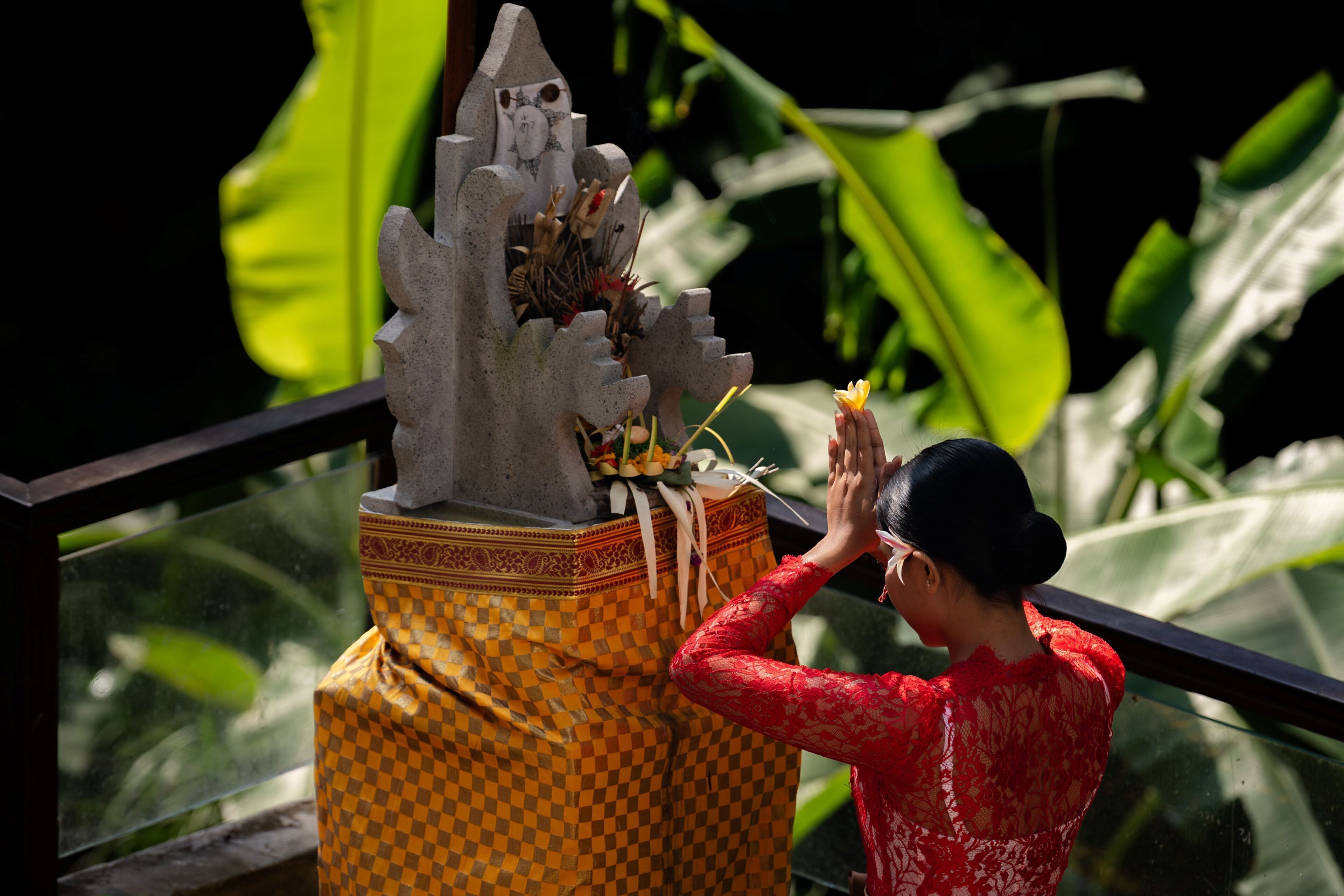 A person in a red dress performing a ritual at a stone statue with a checkered cloth and flowers.