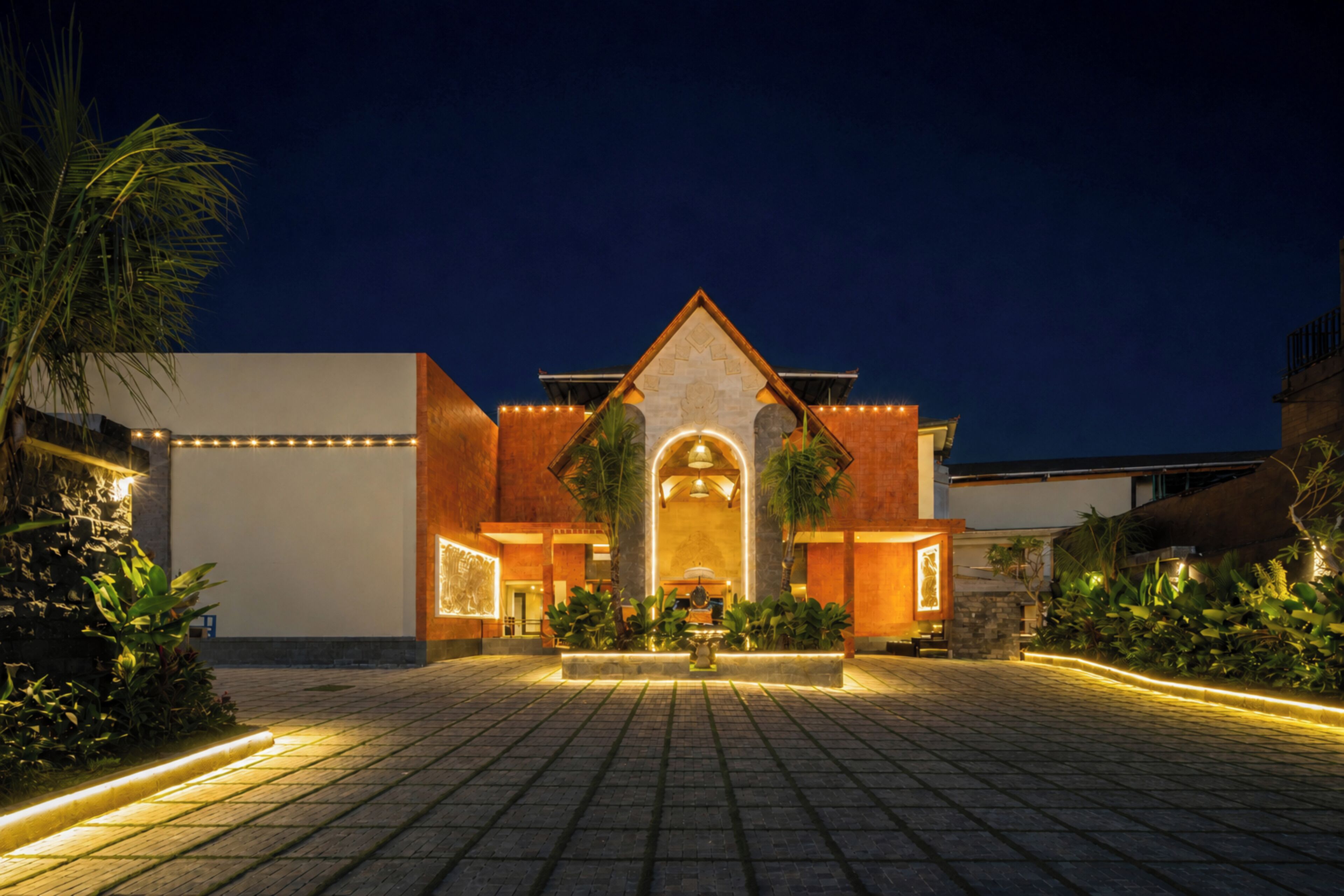 A modern building with a central entrance, surrounded by a paved area with plants and lighting.